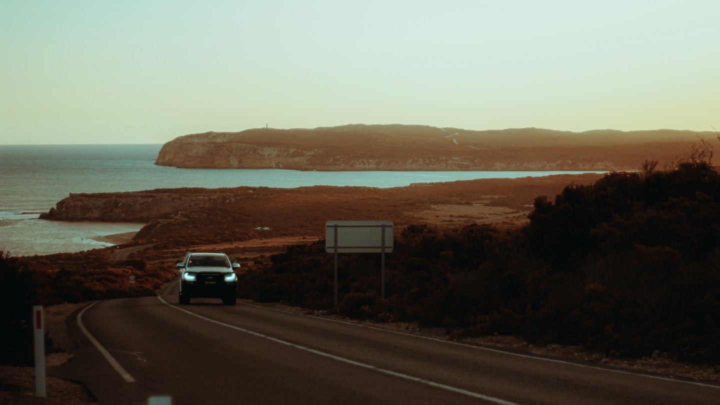 Silver SUV drives on a winding coastal road at sunset.